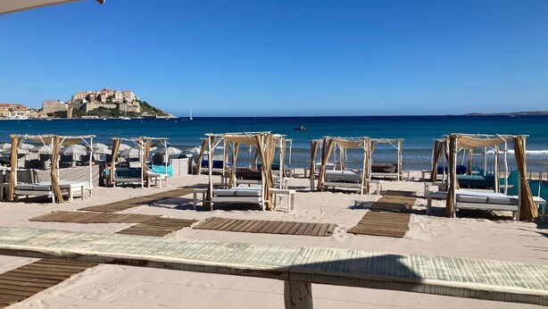 A beach with wooden deck chairs and canopies, a clear blue sky, and a distant view of a castle on a hill. - At sandra&fred your vacation in the dream of our cokon (Calvi)