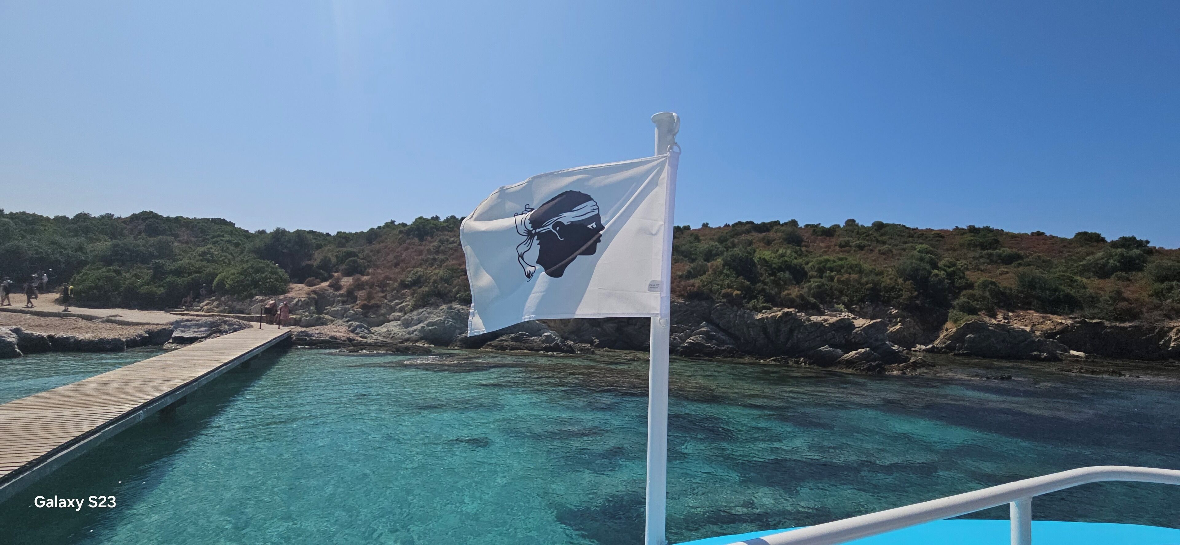 A clear blue sea with a wooden dock extending into it, a flag with a black silhouette, and rocky terrain in the background.