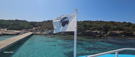 A clear blue sea with a wooden dock extending into it, a flag with a black silhouette, and rocky terrain in the background.