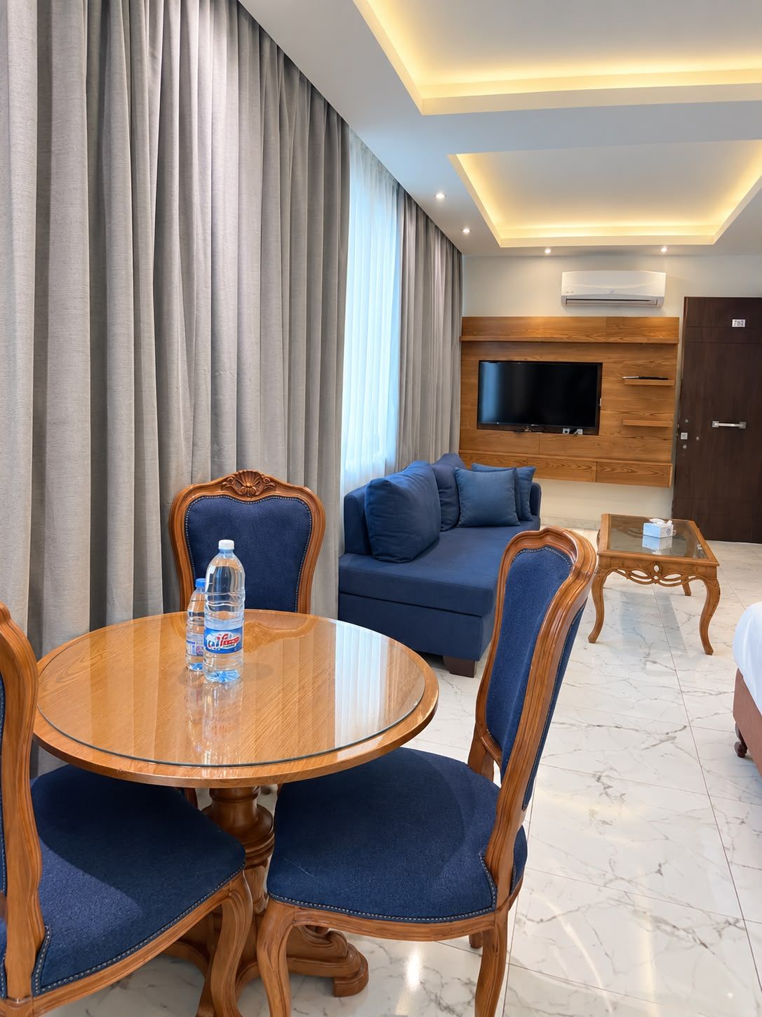 A hotel room with a round wooden dining table, blue upholstered chairs, a blue sofa, a flat-screen TV mounted on a wooden wall, and a marble floor.