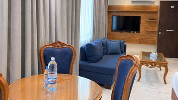 A hotel room with a round wooden dining table, blue upholstered chairs, a blue sofa, a flat-screen TV mounted on a wooden wall, and a marble floor.