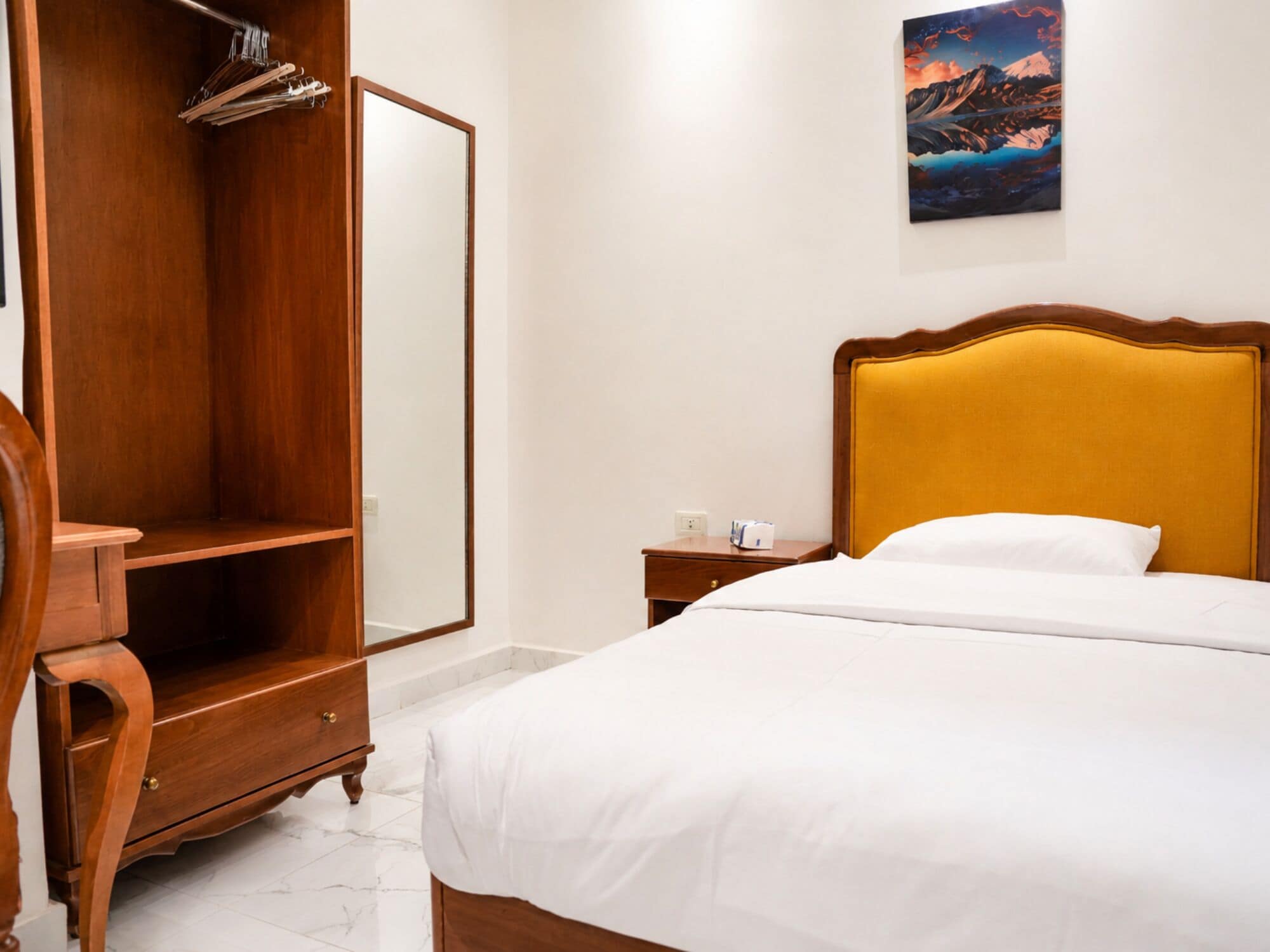 Habitación de hotel con cama, armario de madera, mesita de noche y un cuadro en la pared.