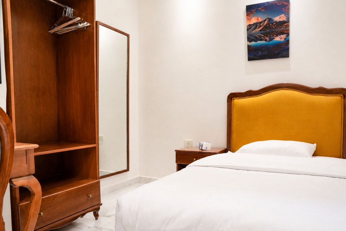A hotel room with a bed, a wooden wardrobe, a nightstand, and a painting on the wall.