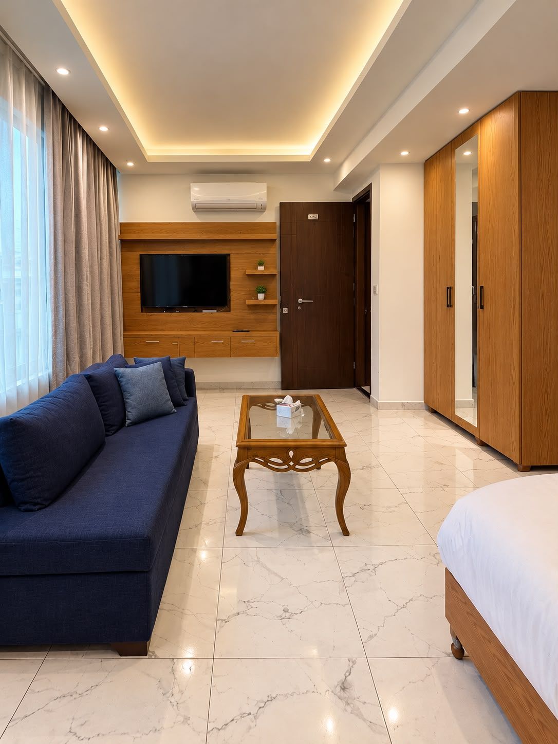 A modern hotel room with a sofa, a glass coffee table, a bed, a TV, and a wardrobe.