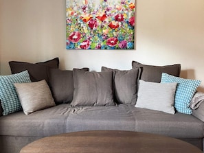 A grey sofa with multiple cushions, a wooden coffee table, and a colorful floral painting on the wall.