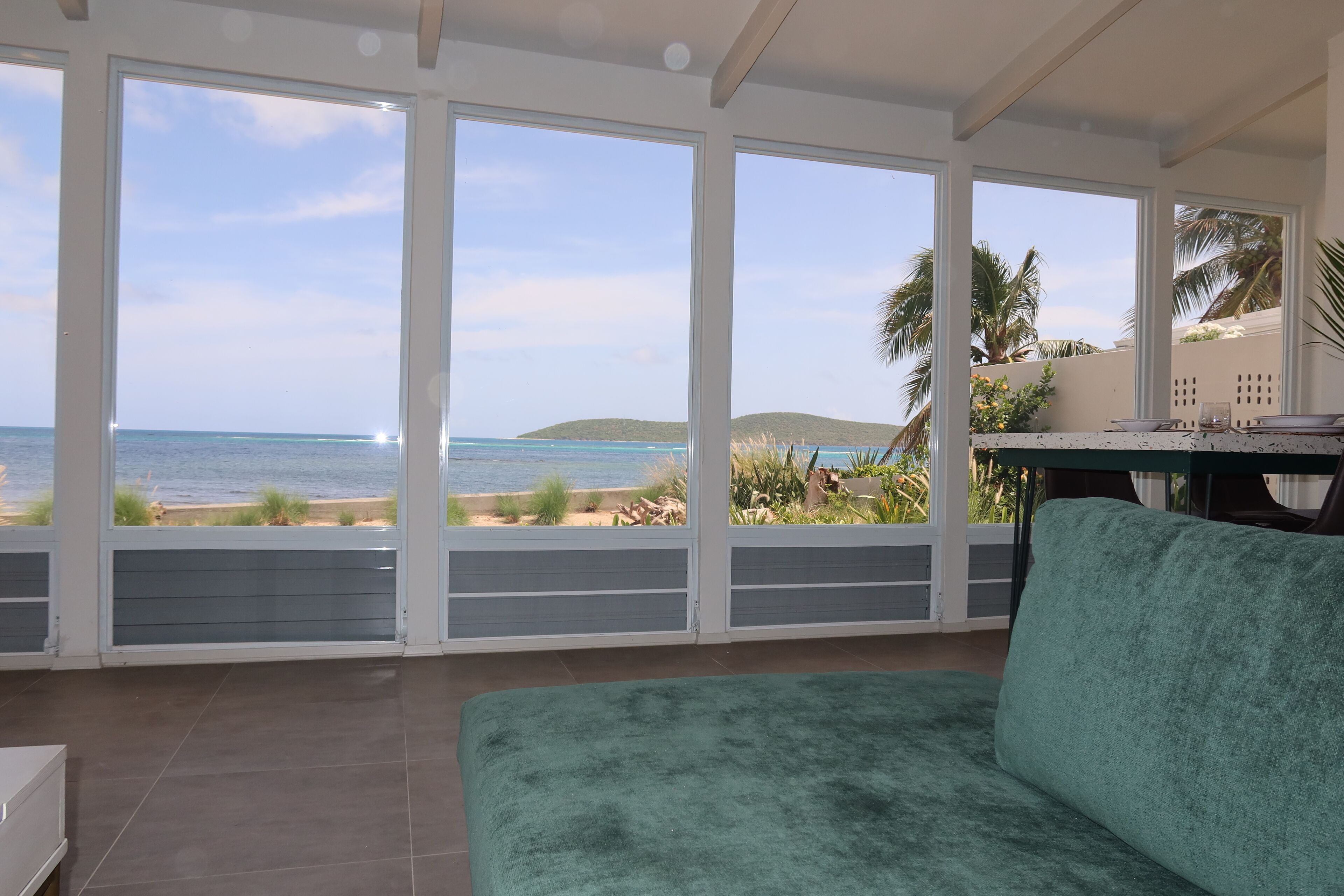 A room with large windows overlooking a beach, a green sofa, and a table with a chair.