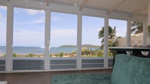 A room with large windows overlooking a beach, a green sofa, and a table with a chair. - Blu-Entire waterfront house (Christiansted)