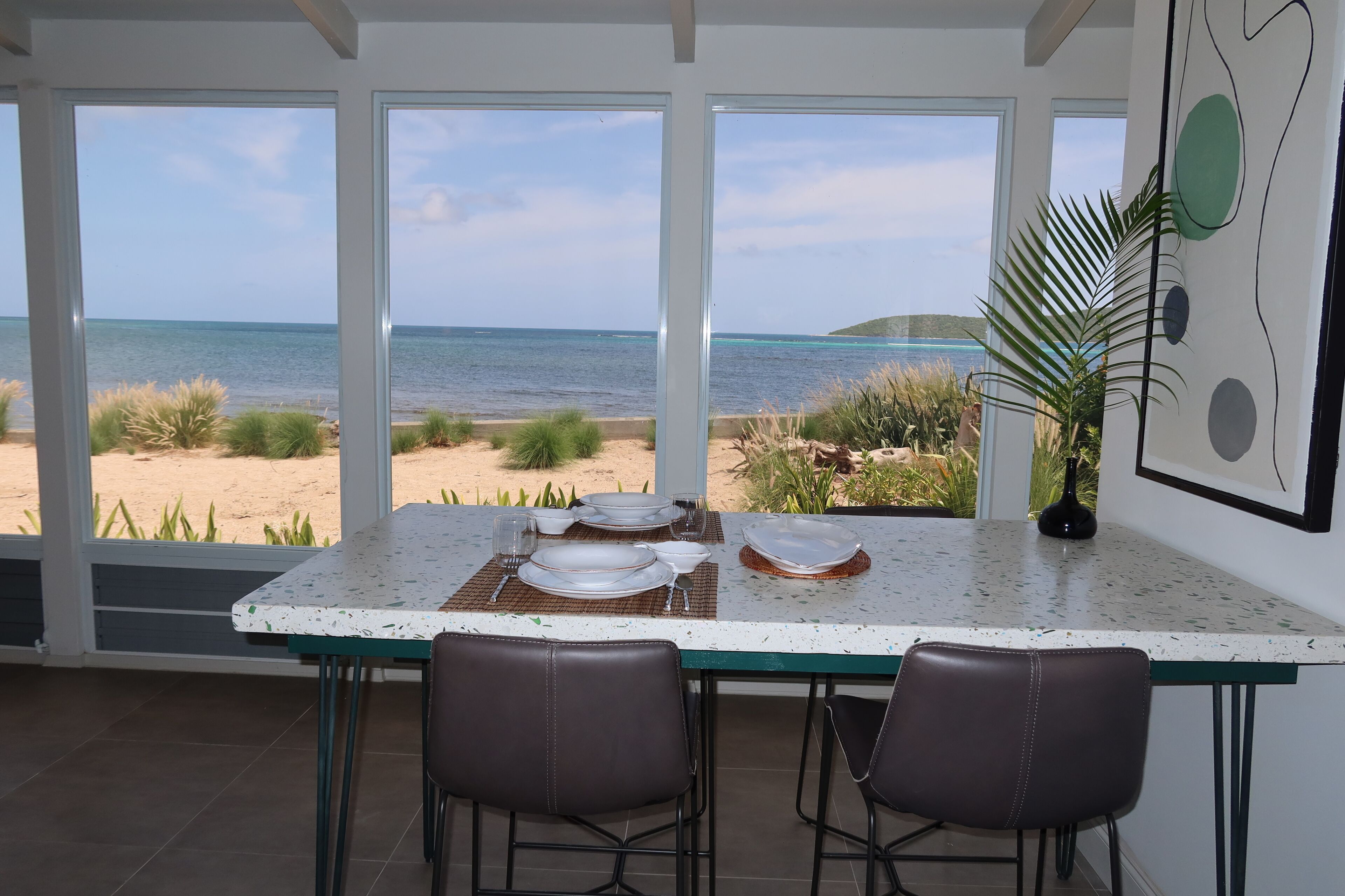 A dining area with a view of the beach and ocean, featuring a table set for two, two chairs, and a decorative plant.