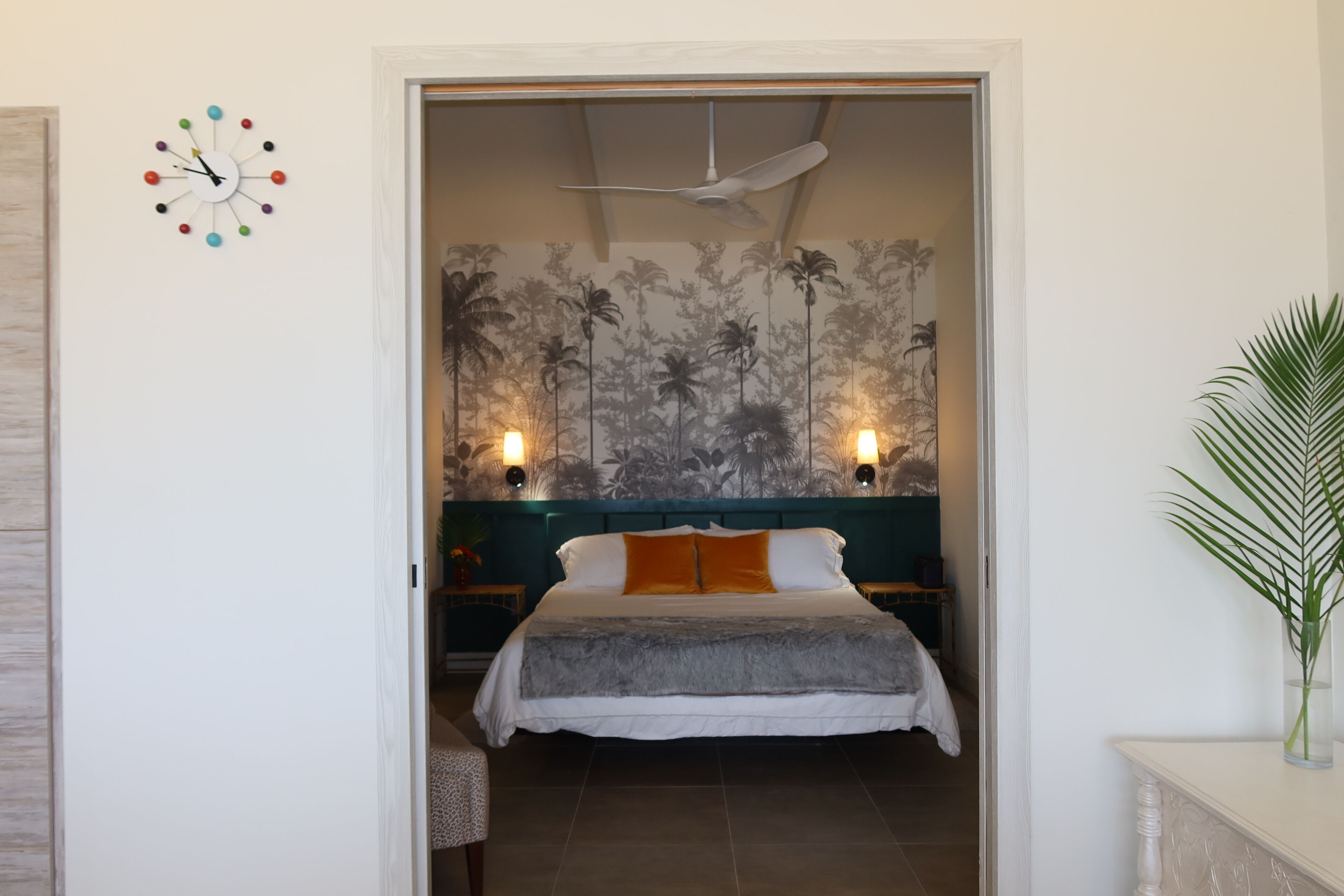 A bedroom with a bed, a ceiling fan, a wall clock, and a tropical wallpaper.