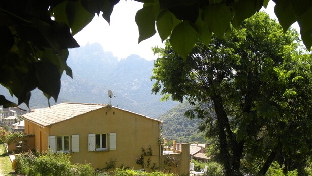 Un bâtiment jaune, doté d’un toit en pente, entouré de verdure et avec une chaîne de montagnes en arrière-plan.