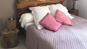 Un lit avec une tête de lit en bois, des oreillers blancs et roses, un petit tabouret en bois et une lampe sur une table de chevet.