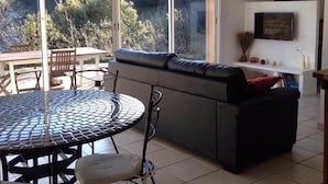 Un salon moderne avec un canapé, une table ronde et des chaises. Un téléviseur est fixé au mur et une fenêtre offre une vue sur les arbres.