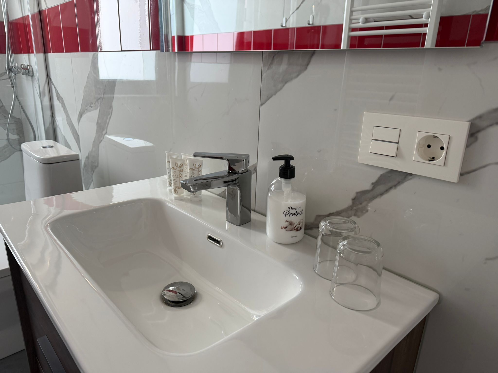 A bathroom with a white sink, a mirror, a red and white tiled backsplash, a handheld showerhead, a bottle of hand soap, and a glass.