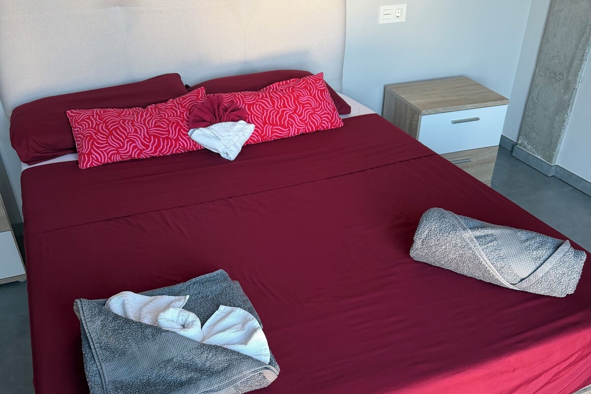 A neatly made bed with a red bedspread, two pillows, and folded towels.