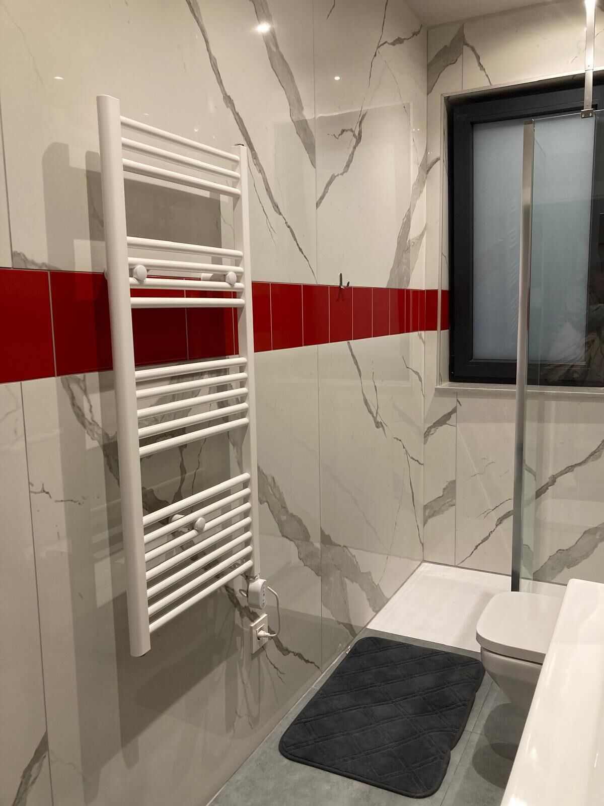 A bathroom with a white towel rack, a red tile accent, a black mat, and a toilet.