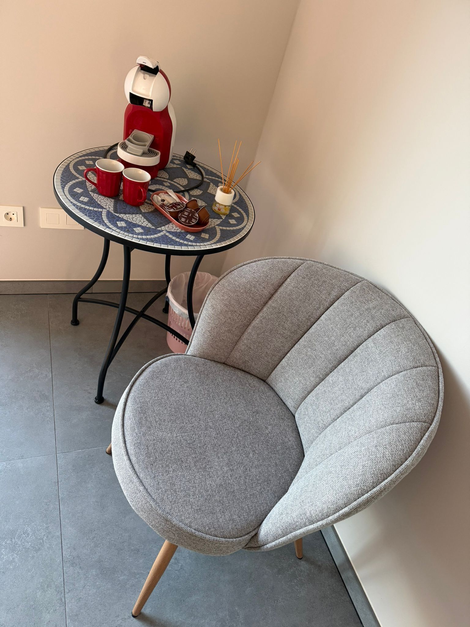 A grey upholstered chair with wooden legs, a small round table with a blue and white mosaic top, and a red and white coffee machine.