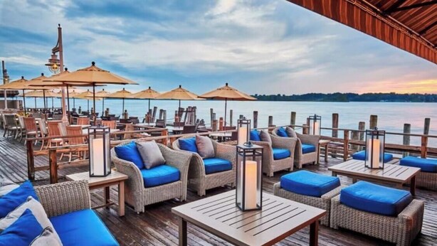 A waterfront deck with wicker furniture, blue cushions, and wooden tables.