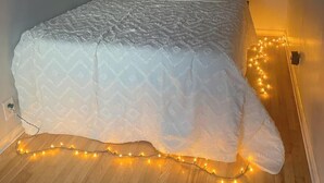 A bed with a white quilt and pillows, a wooden floor, and a string of lights along the wall.
