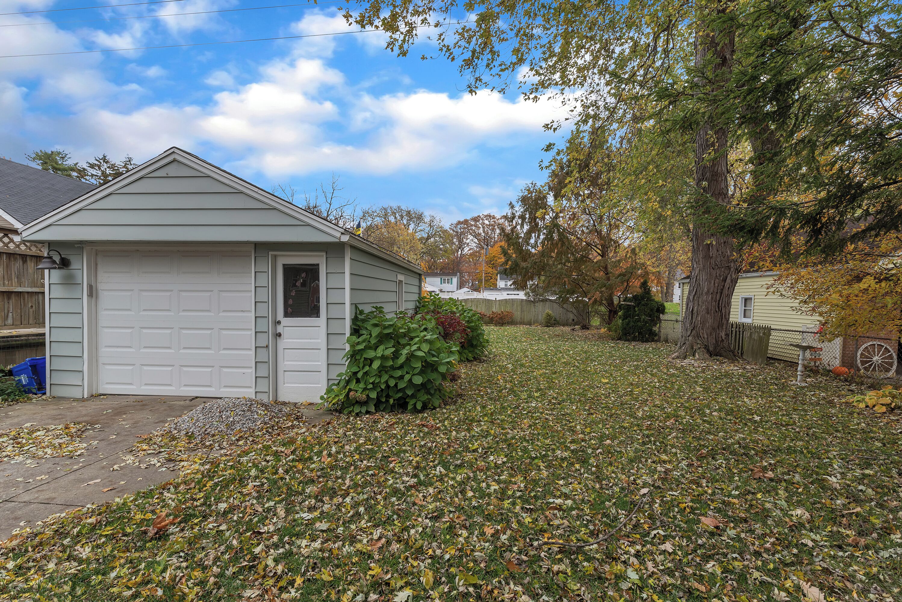 Un garage attenant à une maison, avec une porte blanche, entouré d’une pelouse recouverte de feuilles mortes.