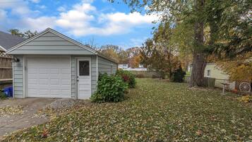 Un garage attenant à une maison, avec une porte blanche, entouré d’une pelouse recouverte de feuilles mortes.