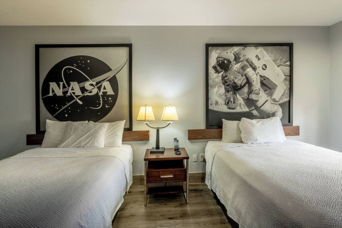 A hotel room with two beds, a nightstand with lamps, NASA-themed wall art, and a wooden floor.