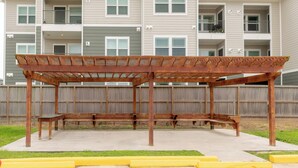 A wooden pergola structure with benches in an outdoor area, surrounded by a wooden fence and a building with multiple balconies in the background. - Azul Inn Houston Hobby Airport (Houston)