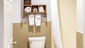 A bathroom with a toilet, a shower, a towel rack, and a shelf with toiletries. - Azul Inn Houston Hobby Airport (Houston)