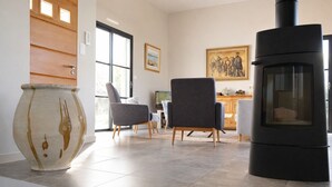 Een moderne woonkamer met een grote, zwarte houtkachel, twee grijze fauteuils, een witte vaas en een houten deur.