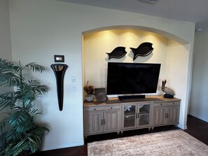 A living room with a flat-screen TV mounted on a wooden cabinet, a wall-mounted light fixture, and a potted plant. - The Salty Marlin in Larry's Harbor POC
 4 Bedrooms w/4 Full Bathrooms (Port O'Connor)