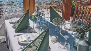 Una mesa lista para una comida con una vista panorámica de la costa, incluyendo una casa y terreno rocoso.