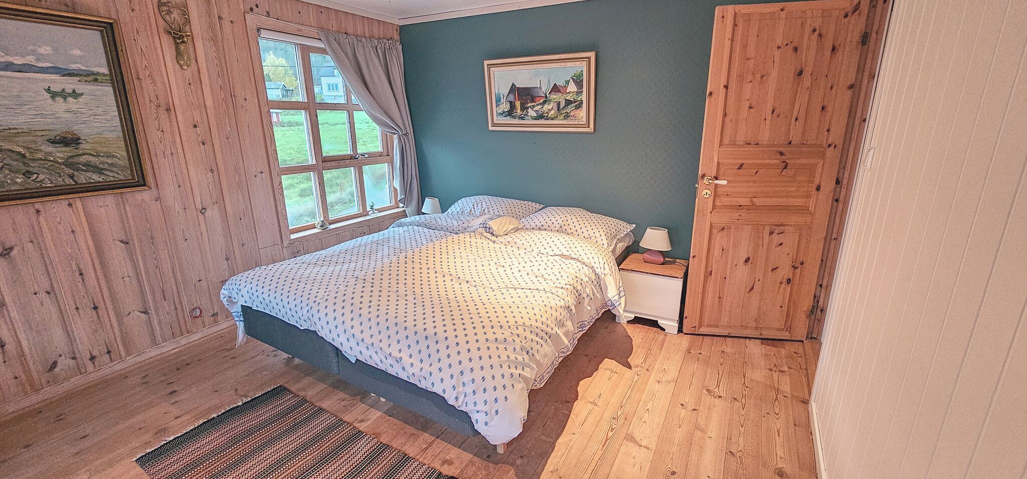 Un dormitorio con cama, mesita de noche, armario de madera, ventana con cortinas y un cuadro en la pared.