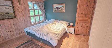 Un dormitorio con cama, mesita de noche, armario de madera, ventana con cortinas y un cuadro en la pared.