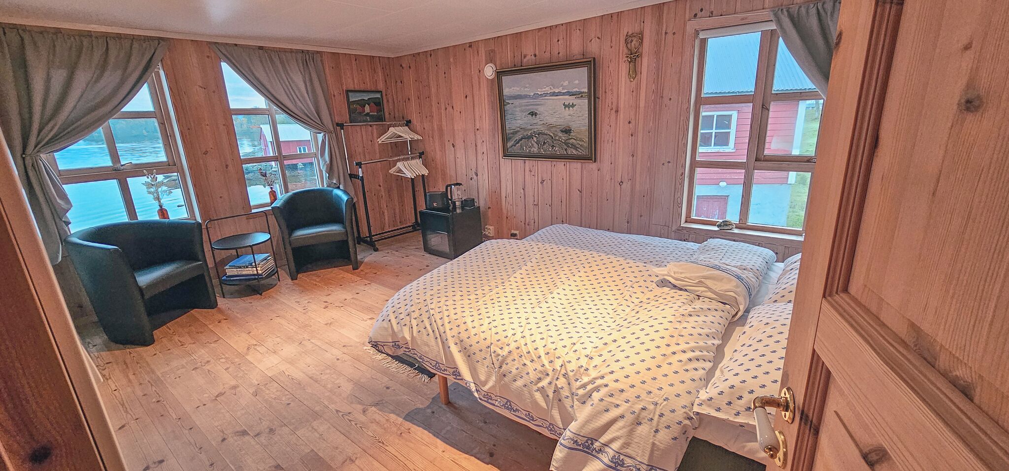 Ein Zimmer mit Holzwänden, einem Bett, zwei Stühlen, einem kleinen Tisch und einem Fenster mit Blick auf ein rotes Gebäude.