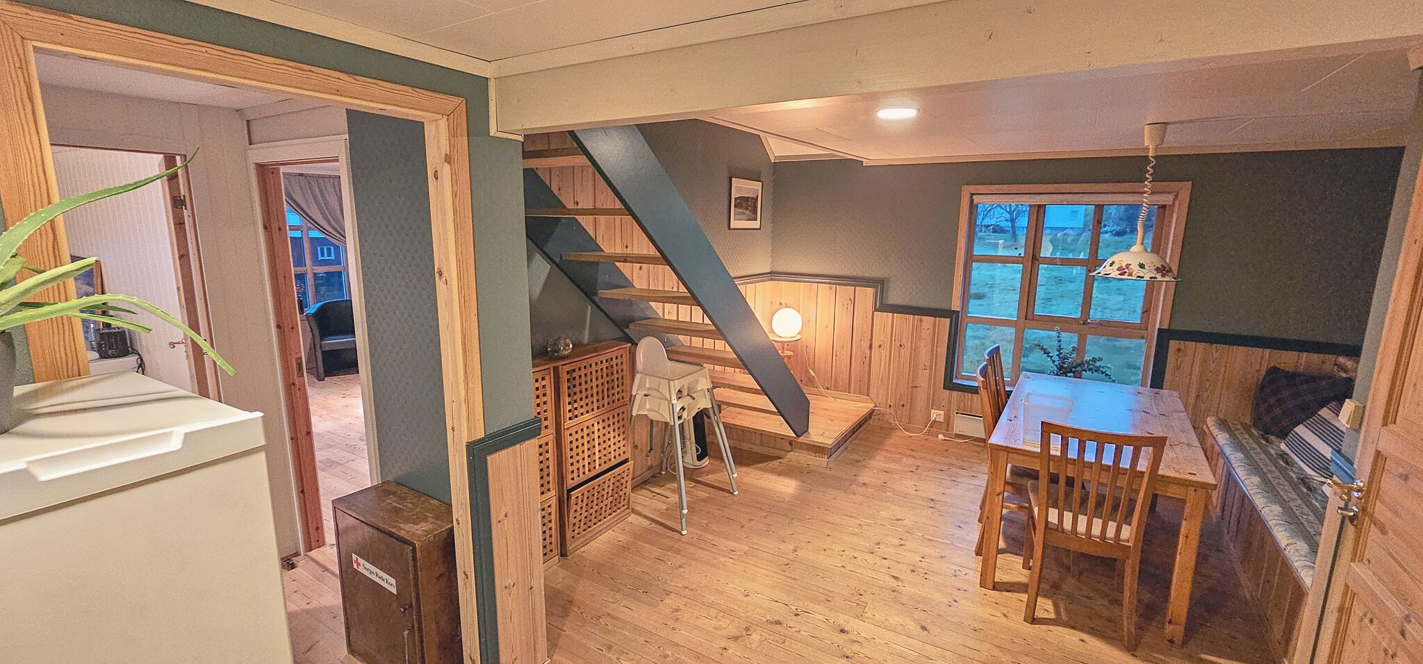 A wooden interior with a dining table, chairs, a staircase, and a view through a window.