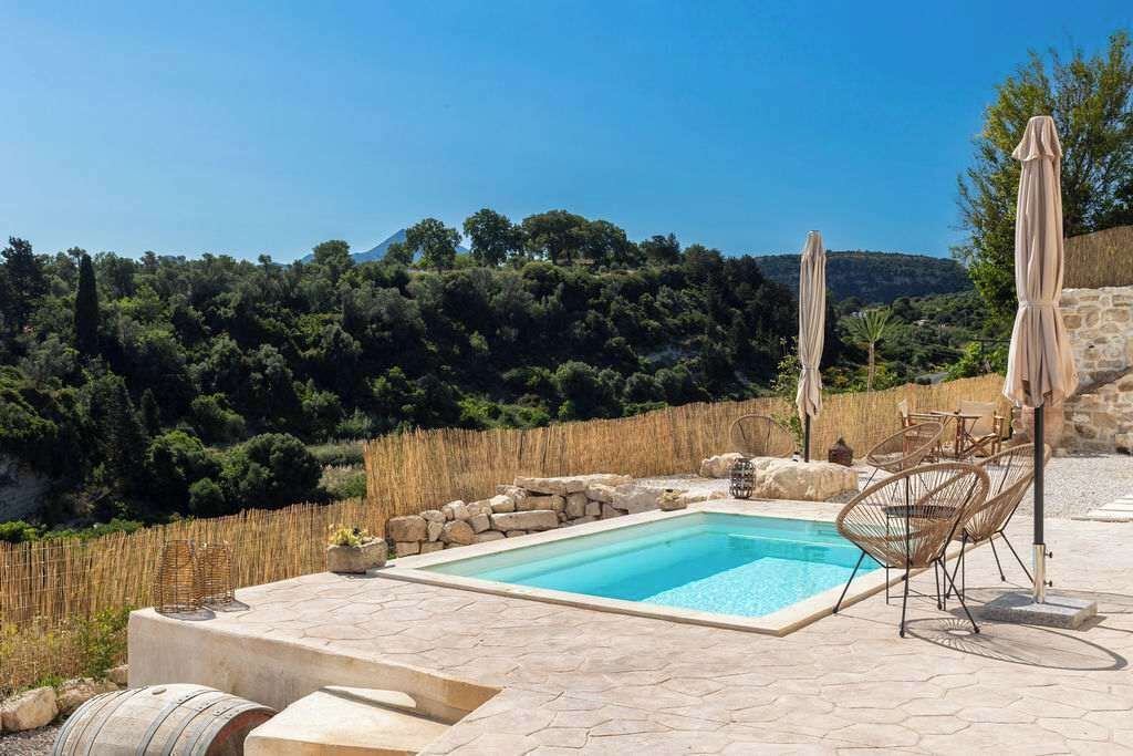 Ein Pool mit Steinfliesen, umgeben von Korbstühlen und Sonnenschirmen, vor einer Kulisse aus Bäumen und Hügeln.