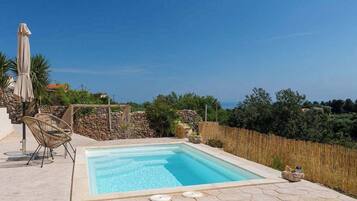 Una piscina con bordo in pietra, sedie in vimini e vista sul mare.