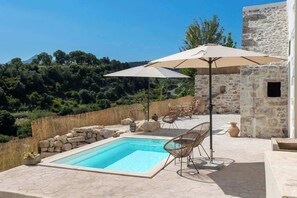 Un espace piscine aménagé avec des chaises longues et un parasol, entouré de murs en pierre et de végétation.