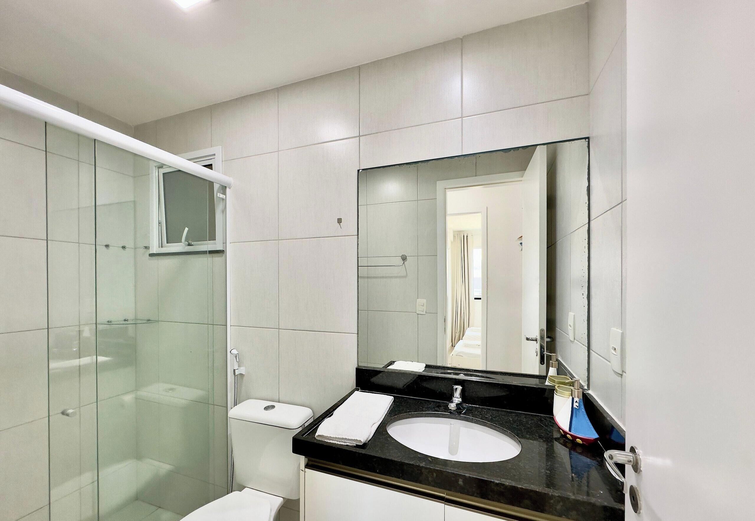 Casa de banho moderna com chuveiro envidraçado, sanita branca, lavatório com espelho e porta-toalhas.