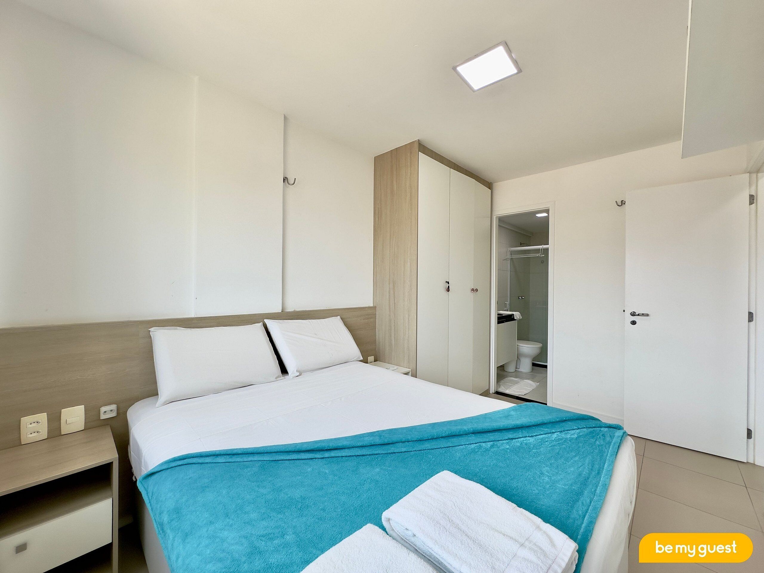 Quarto de hotel com cama, mesa de cabeceira, guarda-roupa e banheiro visível por uma porta aberta.