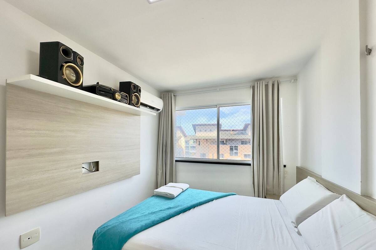 A hotel room with a bed, a window with curtains, a wall-mounted speaker, and a view of buildings outside.