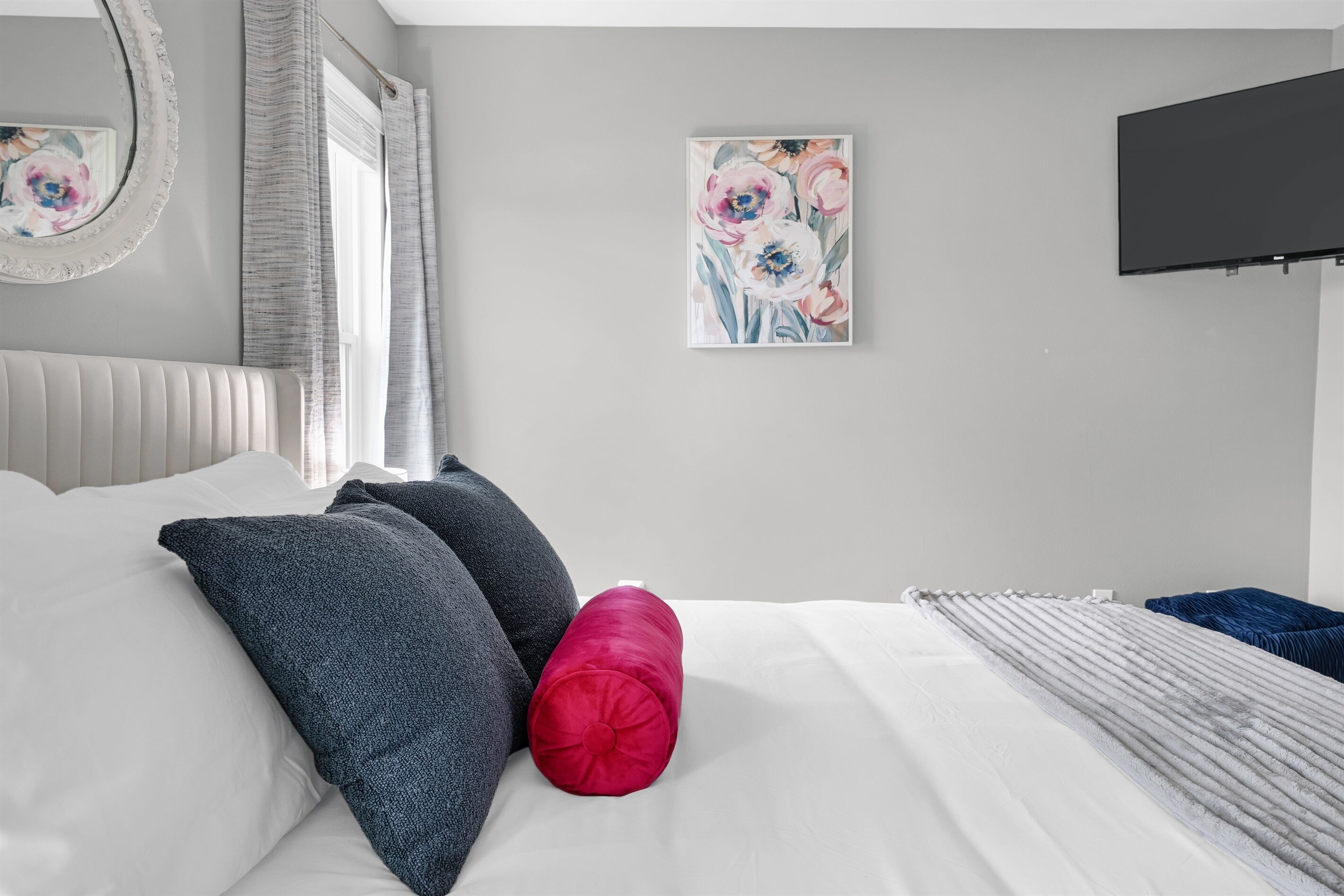 Une chambre à coucher avec un lit, des oreillers, un tableau, une télévision et une fenêtre avec des rideaux.
