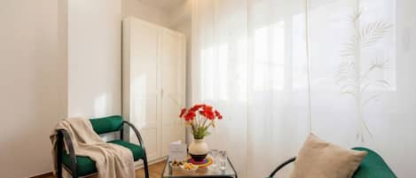 A room with a green sofa, a chair with a blanket, a small table with a vase of flowers, and a white door.