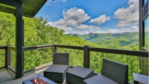 A balcony with wicker furniture and a fire feature, overlooking a mountainous landscape.