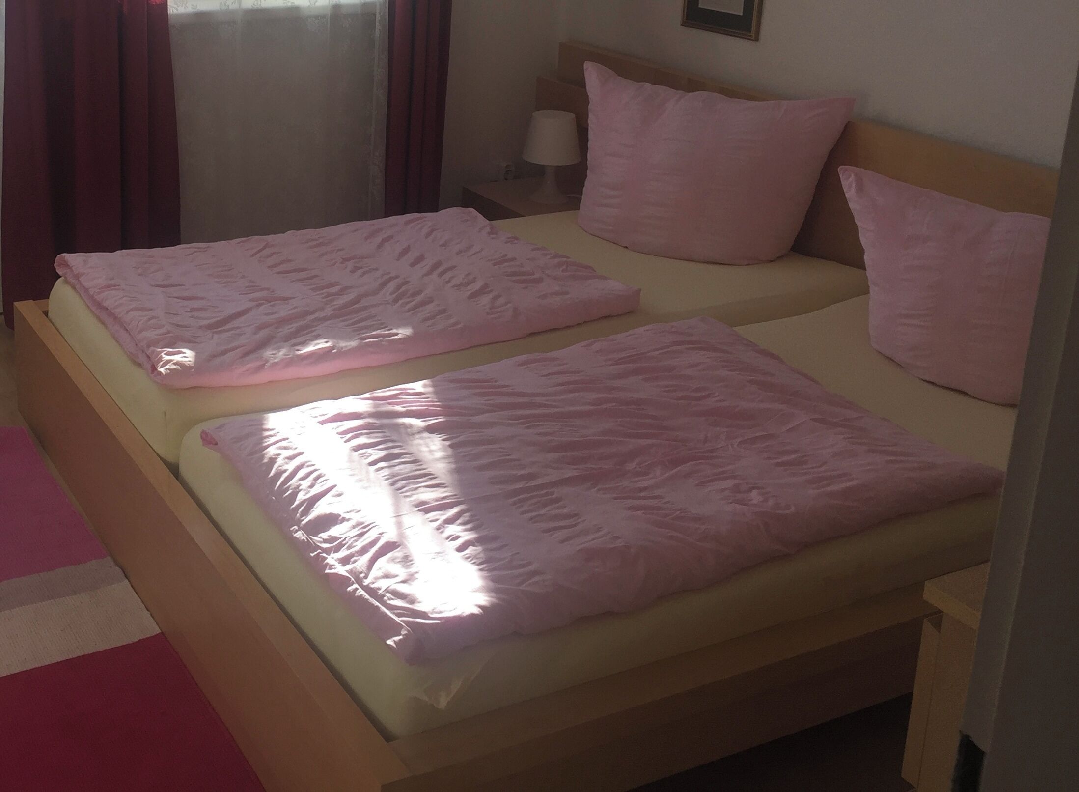 Ein Doppelbett mit rosa Bettwäsche und Kissen, ein Nachttisch mit Lampe und ein Fenster mit Vorhängen.