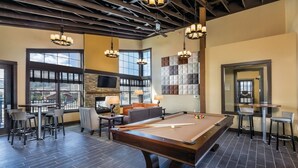 A pool table in a room with a fireplace, large windows, and a bar area.