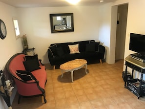 Ein Wohnzimmer mit schwarzem Sofa, einem roten Sessel, einem kleinen weißen Tisch, einem Fernseher auf einem Schrank und einem Bücherregal.