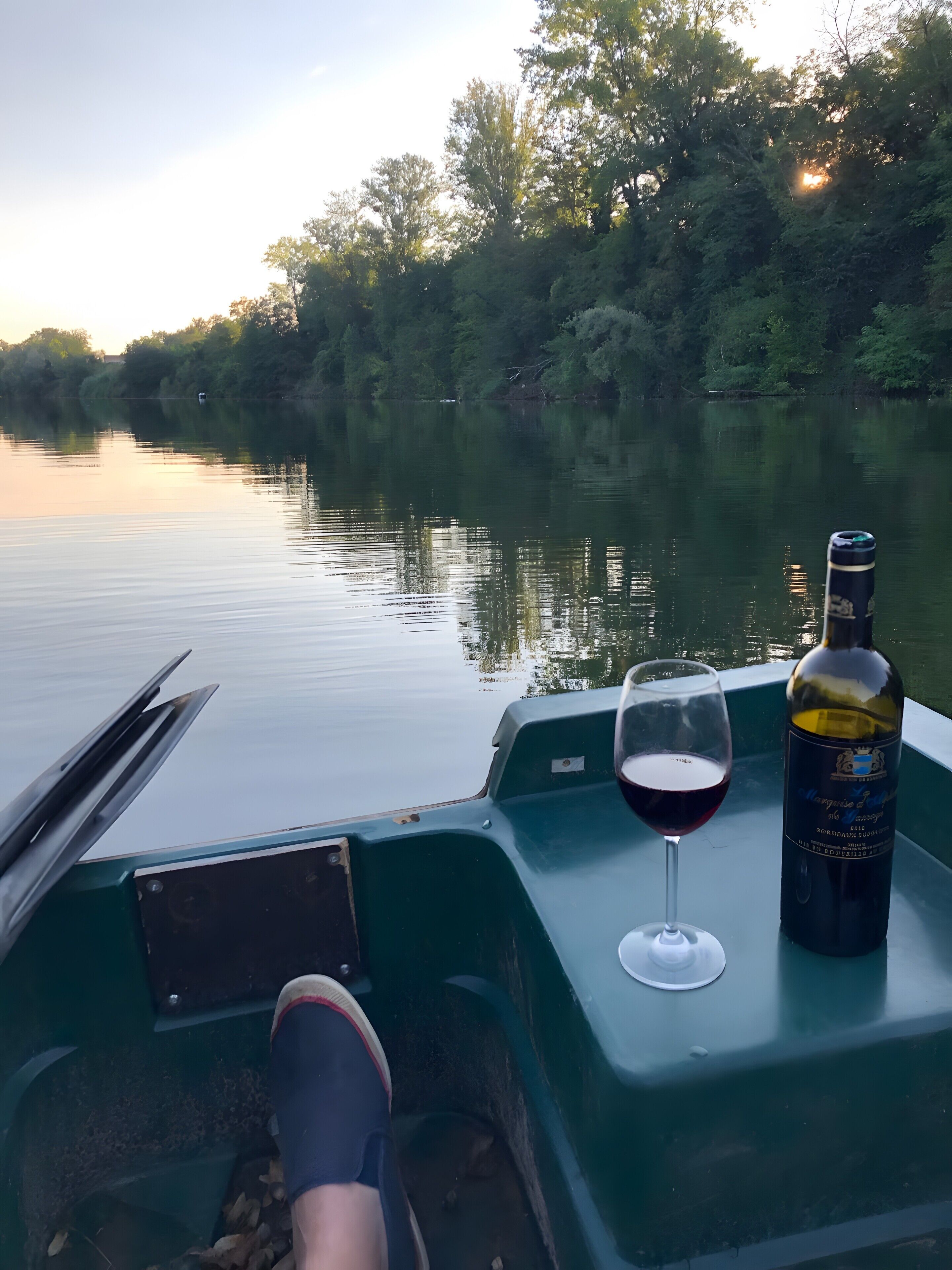 Interior de un barco con una botella de vino y una copa en la cubierta, una persona sentada, y un río tranquilo con árboles al fondo.