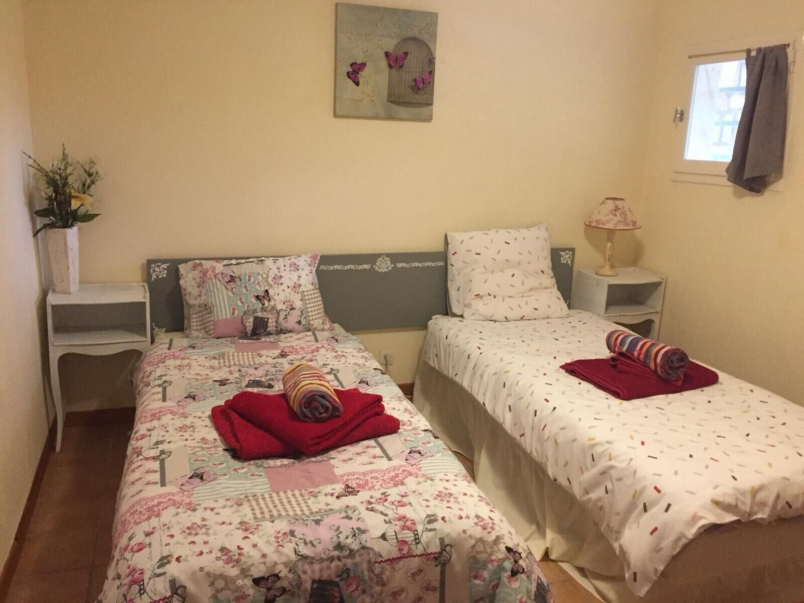 A bedroom with two beds, a bedside table with a lamp, a window with curtains, and a wall-mounted artwork.