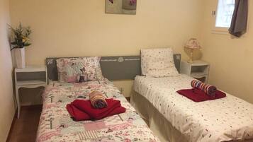 A bedroom with two beds, a bedside table with a lamp, a window with curtains, and a wall-mounted artwork.