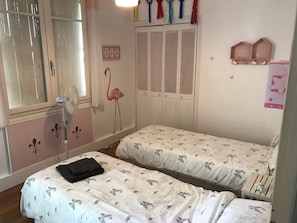 Une chambre avec deux lits, une armoire, une fenêtre avec des stores et des éléments de décoration murale, notamment un flamant rose et une étagère avec des objets en forme d’oiseaux.
